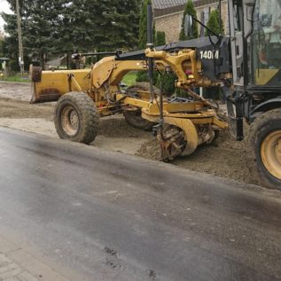 Równiarka profilująca grunt po jego zmieszaniu z dodatkiem hydrofobowym. Sprawny operator pracujący w wąskim pasie pomiędzy pasem z nawierzchnia asfaltową, a krawężnikiem